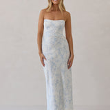 A woman is standing infront of a white backdrop. She's wearing a light blue and white floral print strapless satin dress. The dress is fitted through the bodice and falls nicely over the hips and is tea-length. This dress can be worn as a bridesmaid dress or for any special event. This is the Brodie Dress in Serene.