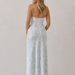 A woman is standing infront of a white backdrop facing the back. She's wearing a light blue and white floral print strapless satin dress. The dress is fitted through the bodice and falls nicely over the hips and is tea-length. This dress can be worn as a bridesmaid dress or for any special event. This is the Brodie Dress in Serene.
