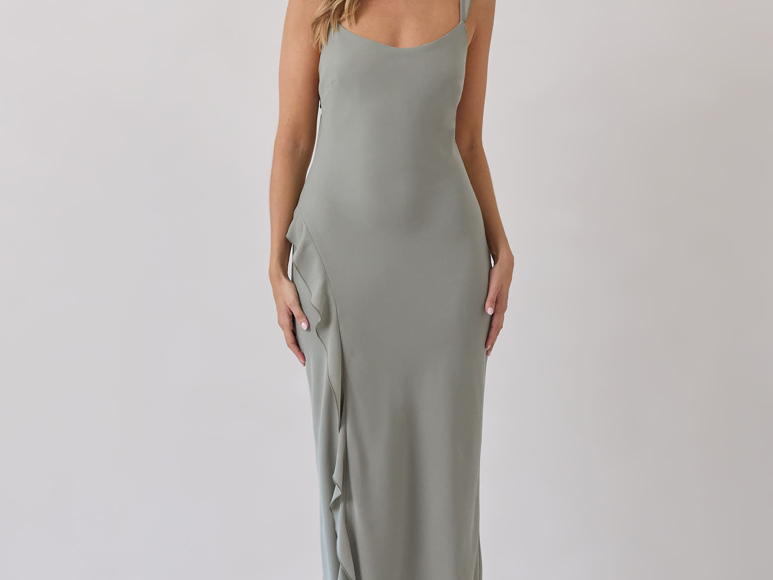 A woman is standing against a white backdrop wearing a sage green bridesmaid dress with an open back. The dress features a slight ruffle detail along the strap and slit of the skirt. This is the Avondale Dress in True Sage.