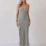 A woman is standing against a white backdrop wearing a sage green bridesmaid dress with an open back. The dress features a slight ruffle detail along the strap and slit of the skirt. This is the Avondale Dress in True Sage.