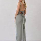 A woman is standing against a white backdrop facing the back wearing a sage green bridesmaid dress with an open back. The dress features a slight ruffle detail along the strap and slit of the skirt. This is the Avondale Dress in True Sage.