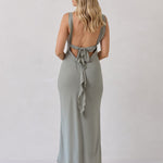 A woman is standing against a white backdrop facing the back wearing a sage green bridesmaid dress with an open back. The dress features a slight ruffle detail along the strap and slit of the skirt. This is the Avondale Dress in True Sage.