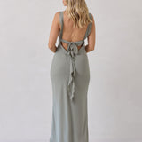 A woman is standing against a white backdrop facing the back wearing a sage green bridesmaid dress with an open back. The dress features a slight ruffle detail along the strap and slit of the skirt. This is the Avondale Dress in True Sage.