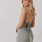 A woman is standing against a white backdrop facing the back wearing a sage green bridesmaid dress with an open back. The dress features a slight ruffle detail along the strap and slit of the skirt. This is the Avondale Dress in True Sage.