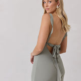 A woman is standing against a white backdrop facing the back wearing a sage green bridesmaid dress with an open back. The dress features a slight ruffle detail along the strap and slit of the skirt. This is the Avondale Dress in True Sage.