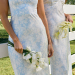 Two women are standing against a fence wearing two different bridesmaid dresses in the same light blue and white floral print. The closest one is wearing a strapless dress and holding a small bouquet of white roses. This is the Brodie Dress in Serene.