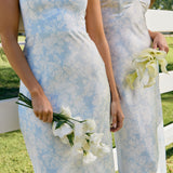 Two women are standing against a fence wearing two different bridesmaid dresses in the same light blue and white floral print. The closest one is wearing a strapless dress and holding a small bouquet of white roses. This is the Brodie Dress in Serene.
