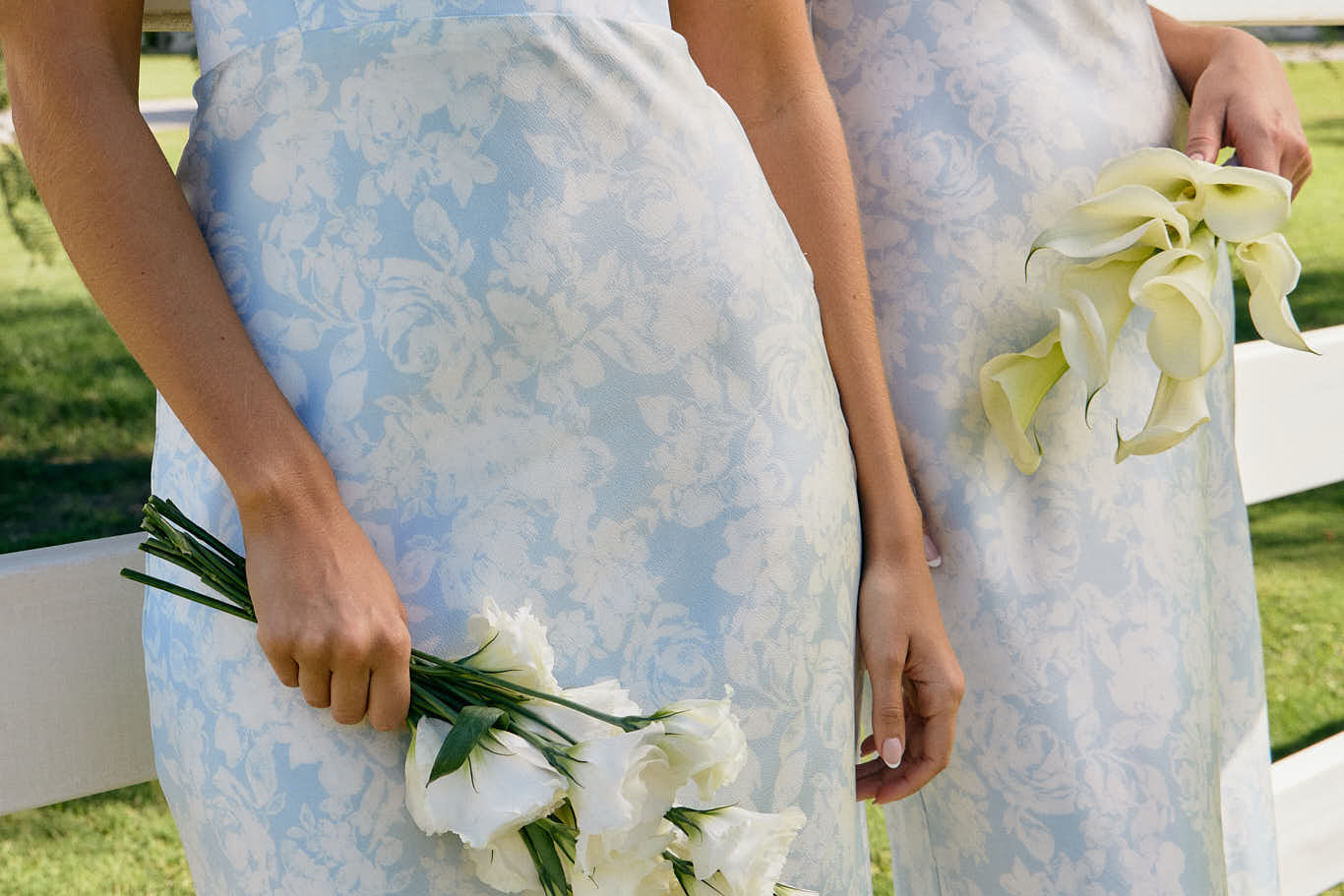 Two women are standing against a fence wearing two different bridesmaid dresses in the same light blue and white floral print. The closest one is wearing a strapless dress and holding a small bouquet of white roses. This is the Brodie Dress in Serene.