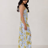 A woman is standing against a white backdrop  wearing a blue and green floral print bridesmaid dress with an open back. The dress features a slight ruffle detail along the strap and slit of the skirt. This is the Avondale Dress in Frankie.