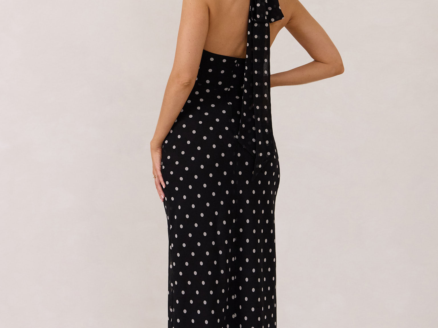 A woman stands against a white backdrop wearing a black and white polka dot dress. The dress has a halter neckline with an optional scarf detail. This is the Archer Dress in Midnight Dot.