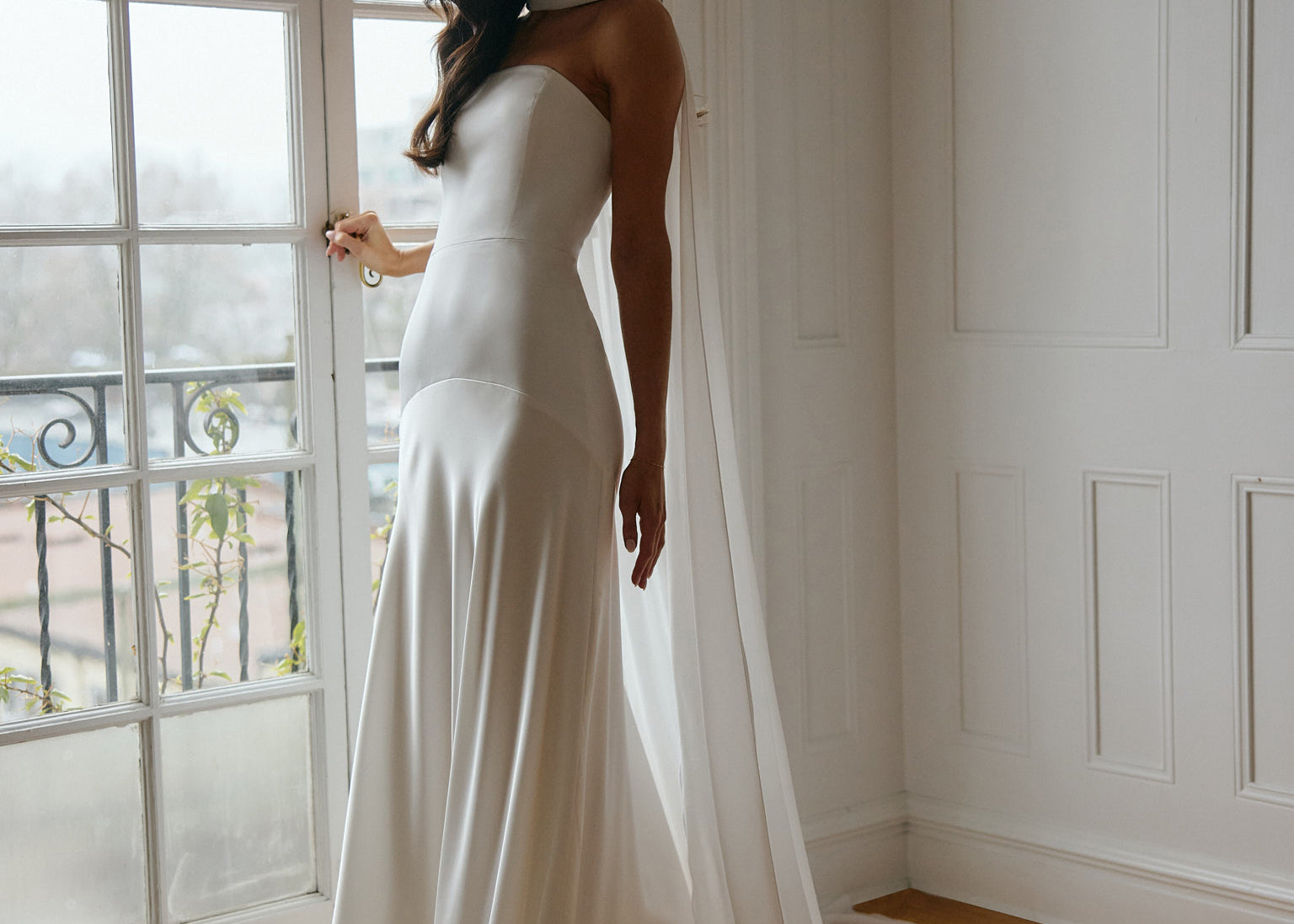 A woman standing in front of a window is wearing a simple white wedding dress with a long, floor length chiffon neck scarf that trails beautifully on the ground. This is the Maeve bridal scarf.