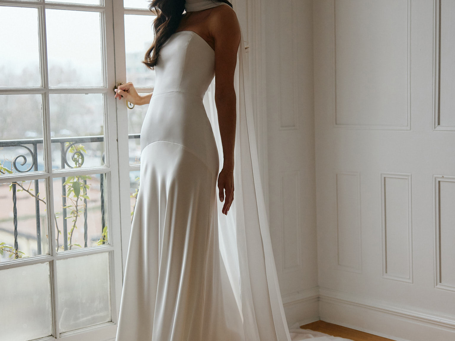 A woman standing in front of a window is wearing a simple white wedding dress with a long, floor length chiffon neck scarf that trails beautifully on the ground. This is the Maeve bridal scarf.