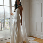 A woman standing in front of a window is wearing a simple white wedding dress with a long, floor length chiffon neck scarf that trails beautifully on the ground. This is the Maeve bridal scarf.
