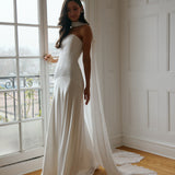 A woman standing in front of a window is wearing a simple white wedding dress with a long, floor length chiffon neck scarf that trails beautifully on the ground. This is the Maeve bridal scarf.