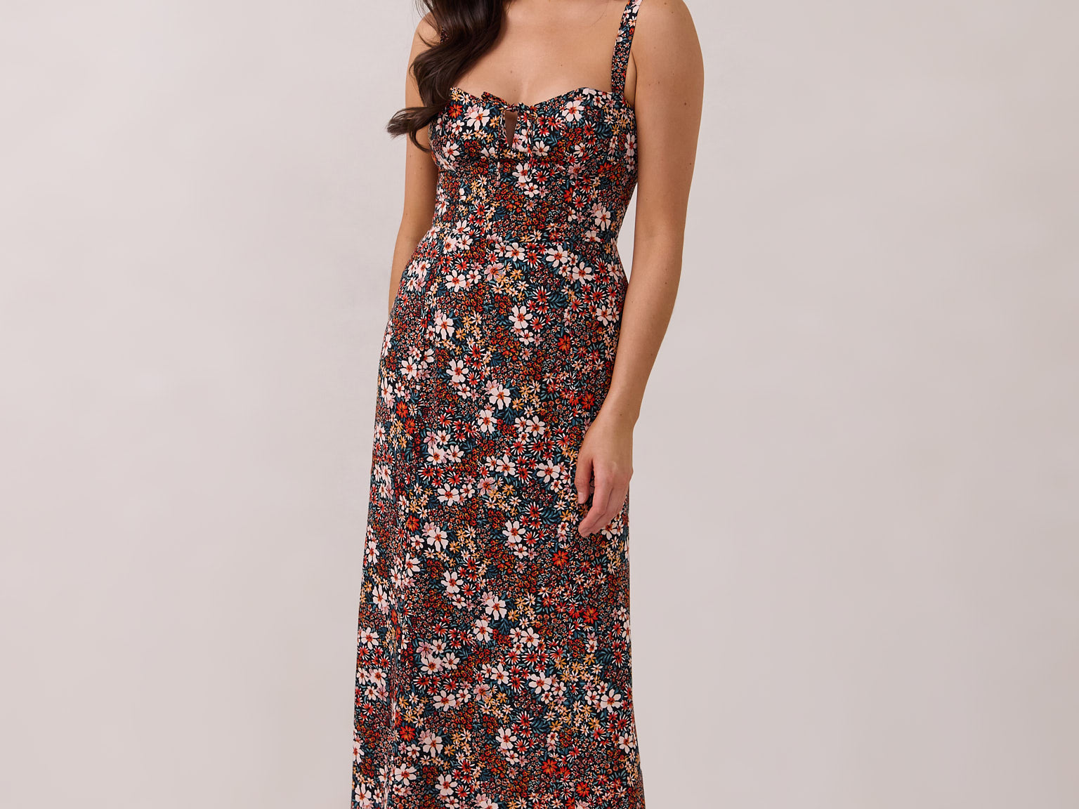 A woman with long dark hair is standing infront of a floral display. She's wearing a floral patterned fitted dress. The dress is fitted through the bodice and has a keyhole detail with a tie at the bust. This spring dress is tea-length and has shirring at the back for added comfort. This is the Decker Dress in Isabel.