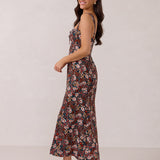 A woman with long dark hair is standing infront of a floral display. She's wearing a floral patterned fitted dress. The dress is fitted through the bodice and has a keyhole detail with a tie at the bust. This spring dress is tea-length and has shirring at the back for added comfort. This is the Decker Dress in Isabel.