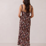 A woman with long dark hair is standing infront of a floral display. She's wearing a floral patterned fitted dress. The dress is fitted through the bodice and has a keyhole detail with a tie at the bust. This spring dress is tea-length and has shirring at the back for added comfort. This is the Decker Dress in Isabel.