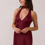 A woman is standing against a white backdrop. She's wearing a burgundy, satin, tea-length bridesmaid dress. The dress has a halter neckline with a scarf detail. The dress is fitted through the bodice and falls nicely over the hips. Perfect to wear as a bridesmaid dress or as a wedding guest dress or any special event.