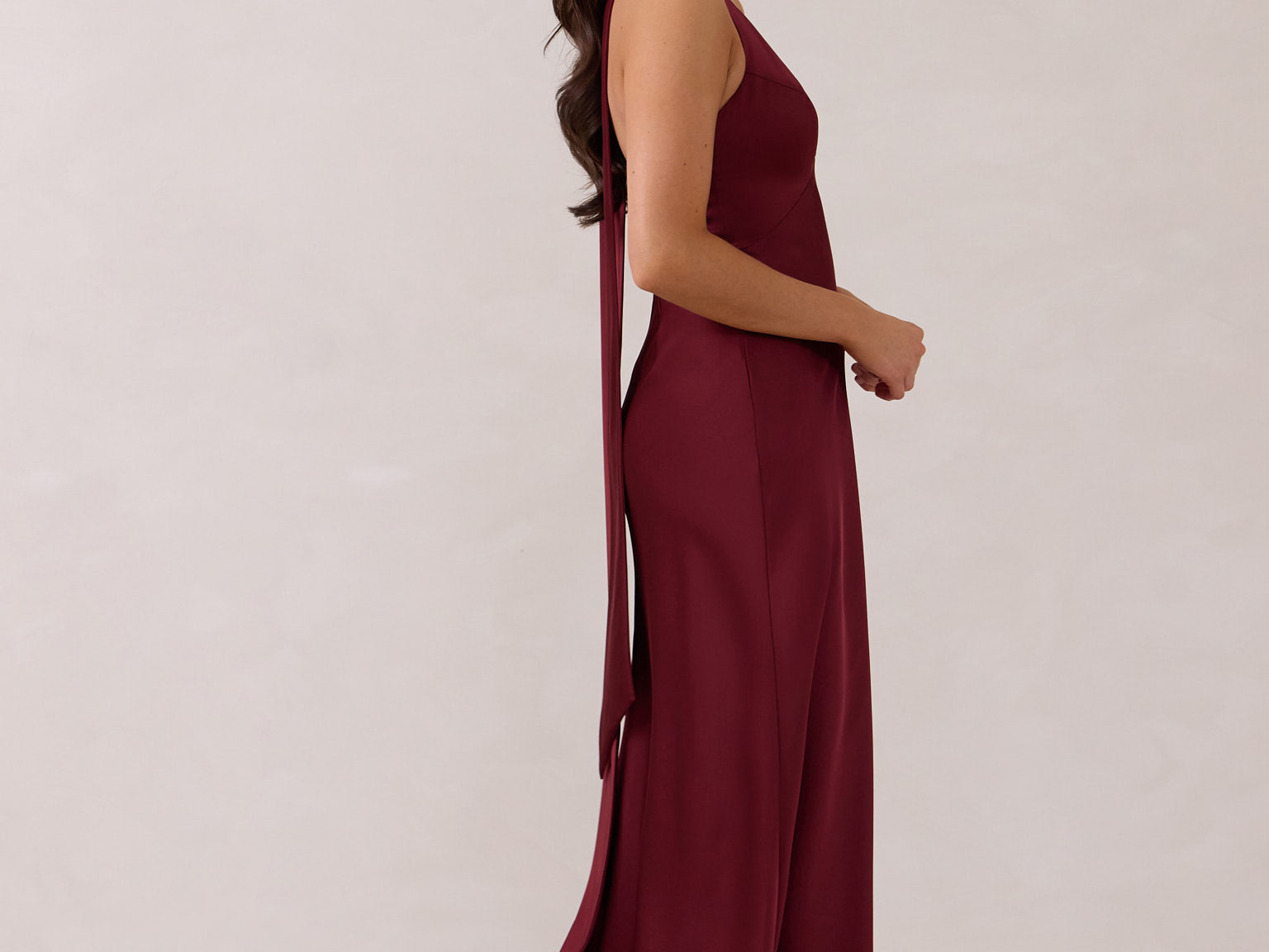 A woman is standing against a white backdrop facing the right. She's wearing a burgundy, satin, tea-length bridesmaid dress. The dress has a halter neckline with a scarf detail. The dress is fitted through the bodice and falls nicely over the hips. Perfect to wear as a bridesmaid dress or as a wedding guest dress or any special event.