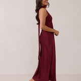 A woman is standing against a white backdrop facing the right. She's wearing a burgundy, satin, tea-length bridesmaid dress. The dress has a halter neckline with a scarf detail. The dress is fitted through the bodice and falls nicely over the hips. Perfect to wear as a bridesmaid dress or as a wedding guest dress or any special event.