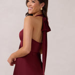 A woman is standing against a white backdrop facing the back. She's wearing a burgundy, satin, tea-length bridesmaid dress. The dress has a halter neckline with a scarf detail. The dress is fitted through the bodice and falls nicely over the hips. Perfect to wear as a bridesmaid dress or as a wedding guest dress or any special event.