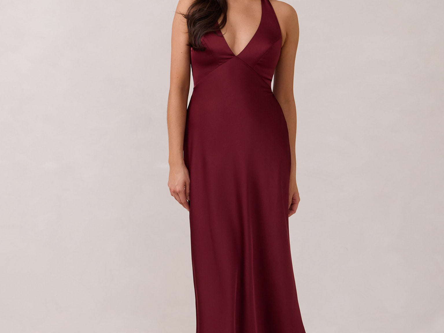 A woman is standing against a white backdrop. She's wearing a burgundy, satin, tea-length bridesmaid dress. The dress has a halter neckline with a scarf detail. The dress is fitted through the bodice and falls nicely over the hips. Perfect to wear as a bridesmaid dress or as a wedding guest dress or any special event.