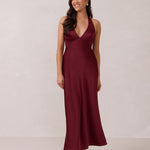 A woman is standing against a white backdrop. She's wearing a burgundy, satin, tea-length bridesmaid dress. The dress has a halter neckline with a scarf detail. The dress is fitted through the bodice and falls nicely over the hips. Perfect to wear as a bridesmaid dress or as a wedding guest dress or any special event.