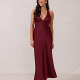 A woman is standing against a white backdrop. She's wearing a burgundy, satin, tea-length bridesmaid dress. The dress has a halter neckline with a scarf detail. The dress is fitted through the bodice and falls nicely over the hips. Perfect to wear as a bridesmaid dress or as a wedding guest dress or any special event.