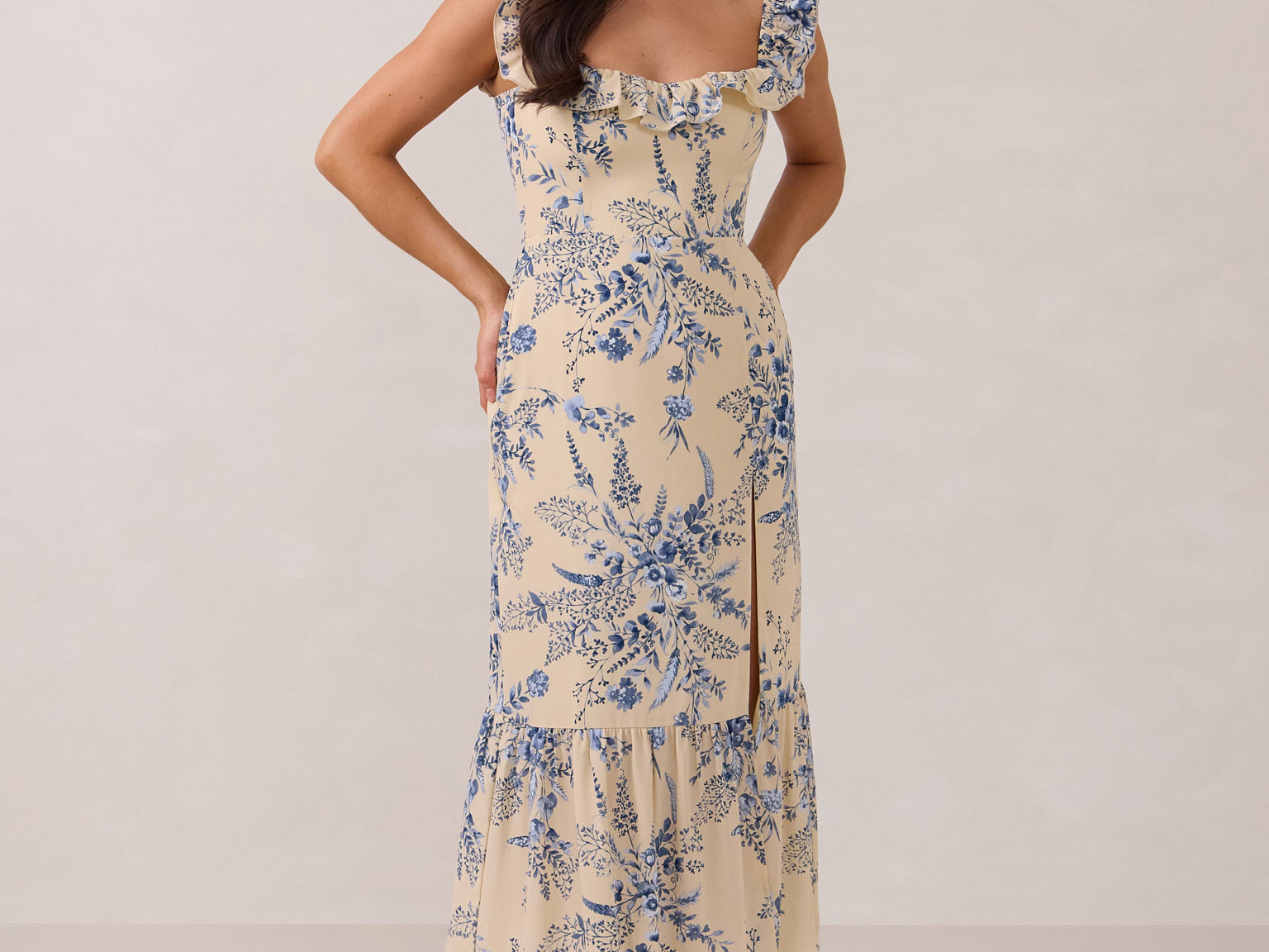 A woman is standing against a white backdrop. She's wearing a tea-length cream dress with a delicate blue floral print. The dress has a ruffle detail around the neckline that can be worn on or off the shoulder. The skirt has a slit up to the thigh and a ruffle detail around the hem. This is the Renfrew Dress in Ayla.