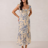 A woman is standing against a white backdrop. She's wearing a tea-length cream dress with a delicate blue floral print. The dress has a ruffle detail around the neckline that can be worn on or off the shoulder. The skirt has a slit up to the thigh and a ruffle detail around the hem. This is the Renfrew Dress in Ayla.