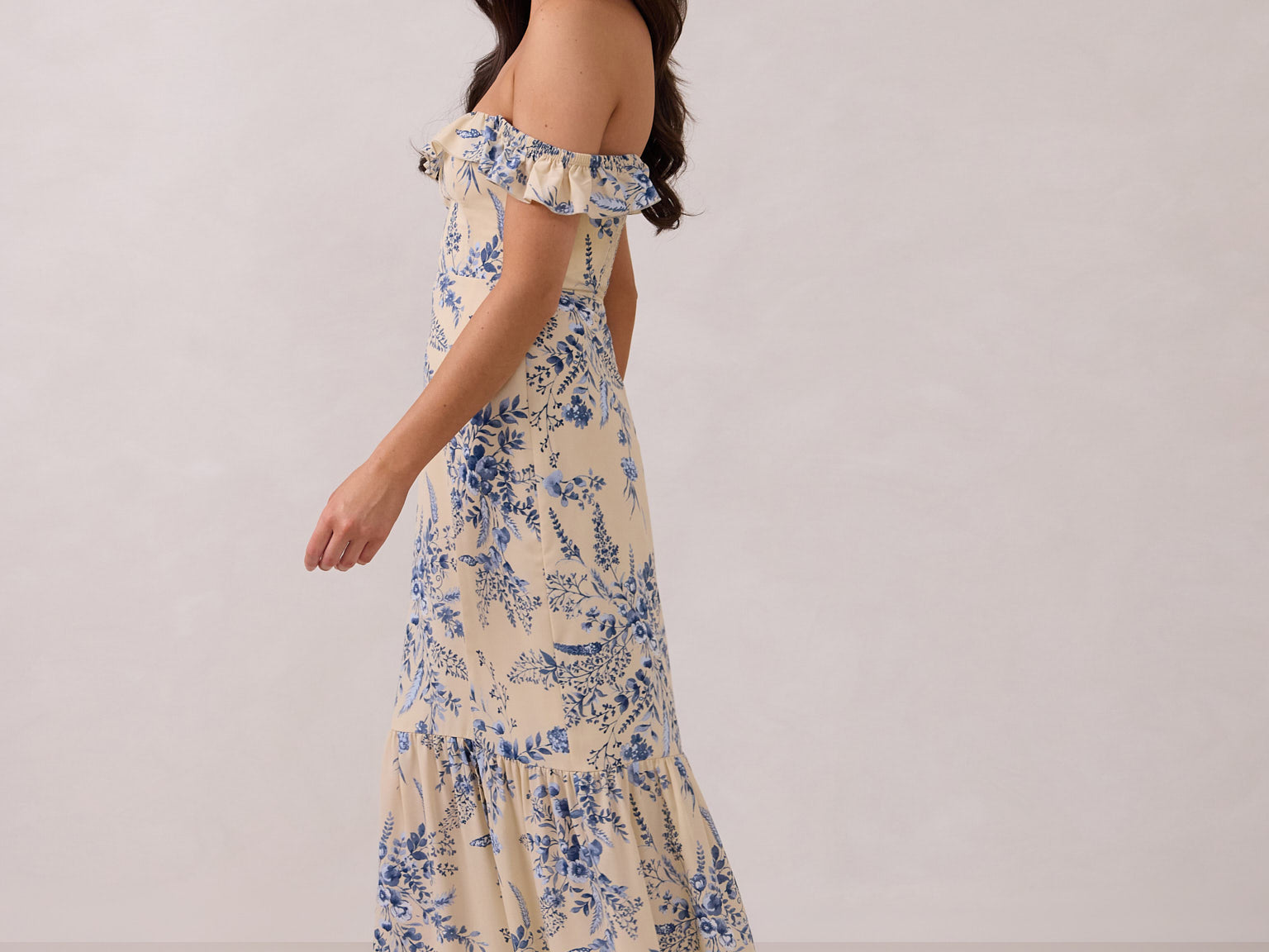 A woman is standing against a white backdrop facing the left. She's wearing a tea-length cream dress with a delicate blue floral print. The dress has a ruffle detail around the neckline that can be worn on or off the shoulder. The skirt has a slit up to the thigh and a ruffle detail around the hem. This is the Renfrew Dress in Ayla.