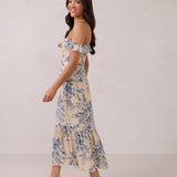 A woman is standing against a white backdrop facing the left. She's wearing a tea-length cream dress with a delicate blue floral print. The dress has a ruffle detail around the neckline that can be worn on or off the shoulder. The skirt has a slit up to the thigh and a ruffle detail around the hem. This is the Renfrew Dress in Ayla.