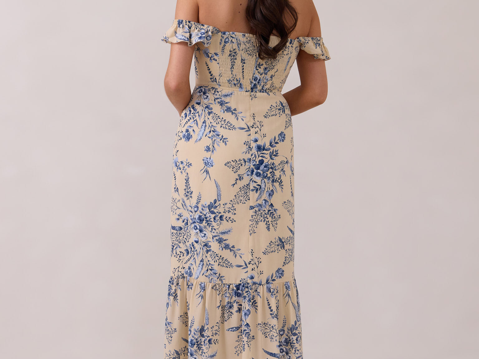 A woman is standing against a white backdrop facing the back. She's wearing a tea-length cream dress with a delicate blue floral print. The dress has a ruffle detail around the neckline that can be worn on or off the shoulder. The skirt has a slit up to the thigh and a ruffle detail around the hem. This is the Renfrew Dress in Ayla.