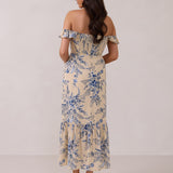A woman is standing against a white backdrop facing the back. She's wearing a tea-length cream dress with a delicate blue floral print. The dress has a ruffle detail around the neckline that can be worn on or off the shoulder. The skirt has a slit up to the thigh and a ruffle detail around the hem. This is the Renfrew Dress in Ayla.