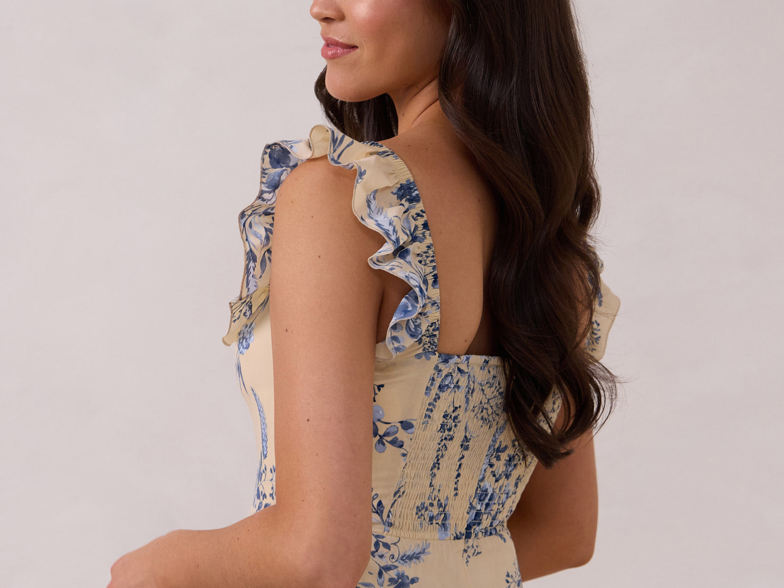A woman is standing against a white backdrop facing the back and looking over her shoulder. She's wearing a tea-length cream dress with a delicate blue floral print. The dress has a ruffle detail around the neckline that can be worn on or off the shoulder. The skirt has a slit up to the thigh and a ruffle detail around the hem. This is the Renfrew Dress in Ayla.