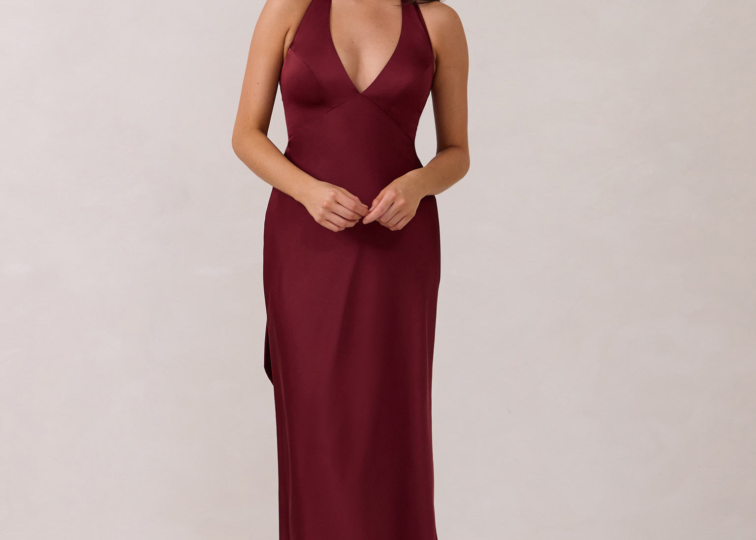 A woman is standing against a white backdrop. She's wearing a burgundy, satin, tea-length bridesmaid dress. The dress has a halter neckline with a scarf detail. The dress is fitted through the bodice and falls nicely over the hips. Perfect to wear as a bridesmaid dress or as a wedding guest dress or any special event.
