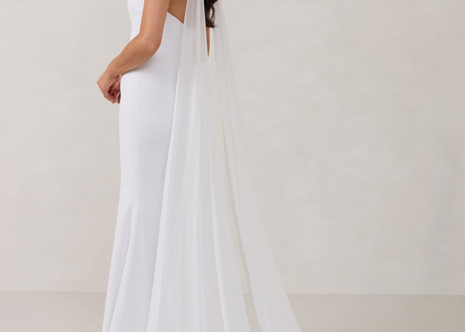 A woman standing in front of a white backdrop is wearing a simple white wedding dress with a long, floor length chiffon neck scarf that trails beautifully on the ground. This is the Maeve bridal scarf.