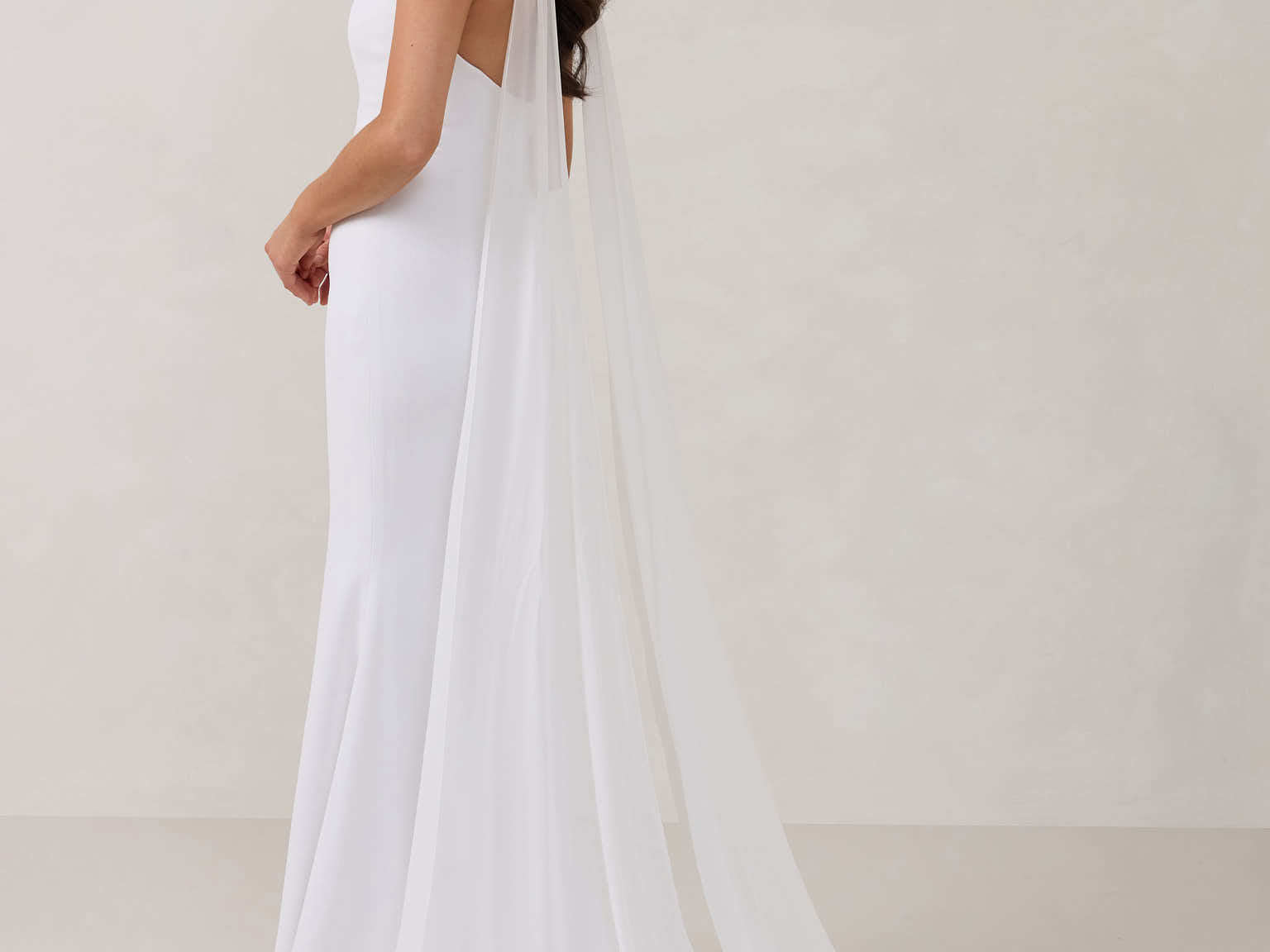 A woman standing in front of a white backdrop is wearing a simple white wedding dress with a long, floor length chiffon neck scarf that trails beautifully on the ground. This is the Maeve bridal scarf.