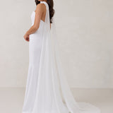 A woman standing in front of a white backdrop is wearing a simple white wedding dress with a long, floor length chiffon neck scarf that trails beautifully on the ground. This is the Maeve bridal scarf.