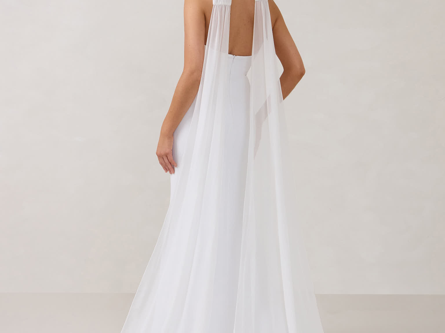 A woman standing facing the back in front of a white backdrop is wearing a simple white wedding dress with a long, floor length chiffon neck scarf that trails beautifully on the ground. This is the Maeve bridal scarf.