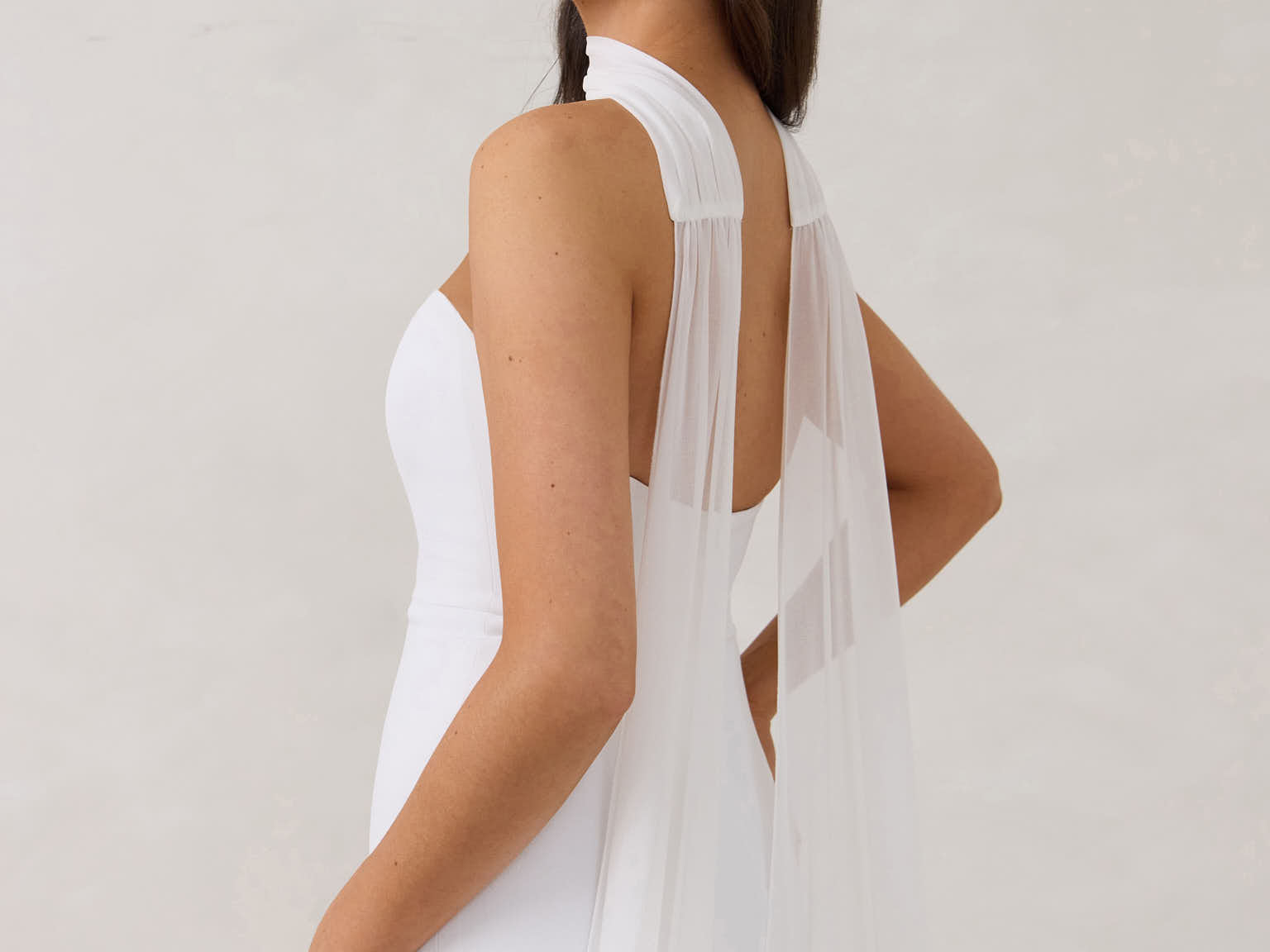 A woman standing in front of a white backdrop is wearing a simple white wedding dress with a long, floor length chiffon neck scarf that trails beautifully on the ground. This is the Maeve bridal scarf.