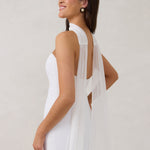 A woman standing in front of a white backdrop is wearing a simple white wedding dress with a long, floor length chiffon neck scarf that trails beautifully on the ground. This is the Maeve bridal scarf.