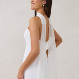 A woman standing in front of a white backdrop is wearing a simple white wedding dress with a long, floor length chiffon neck scarf that trails beautifully on the ground. This is the Maeve bridal scarf.