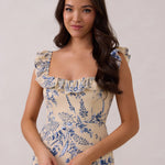 A woman is standing against a white backdrop. She's wearing a tea-length cream dress with a delicate blue floral print. The dress has a ruffle detail around the neckline that can be worn on or off the shoulder. The skirt has a slit up to the thigh and a ruffle detail around the hem. This is the Renfrew Dress in Ayla.