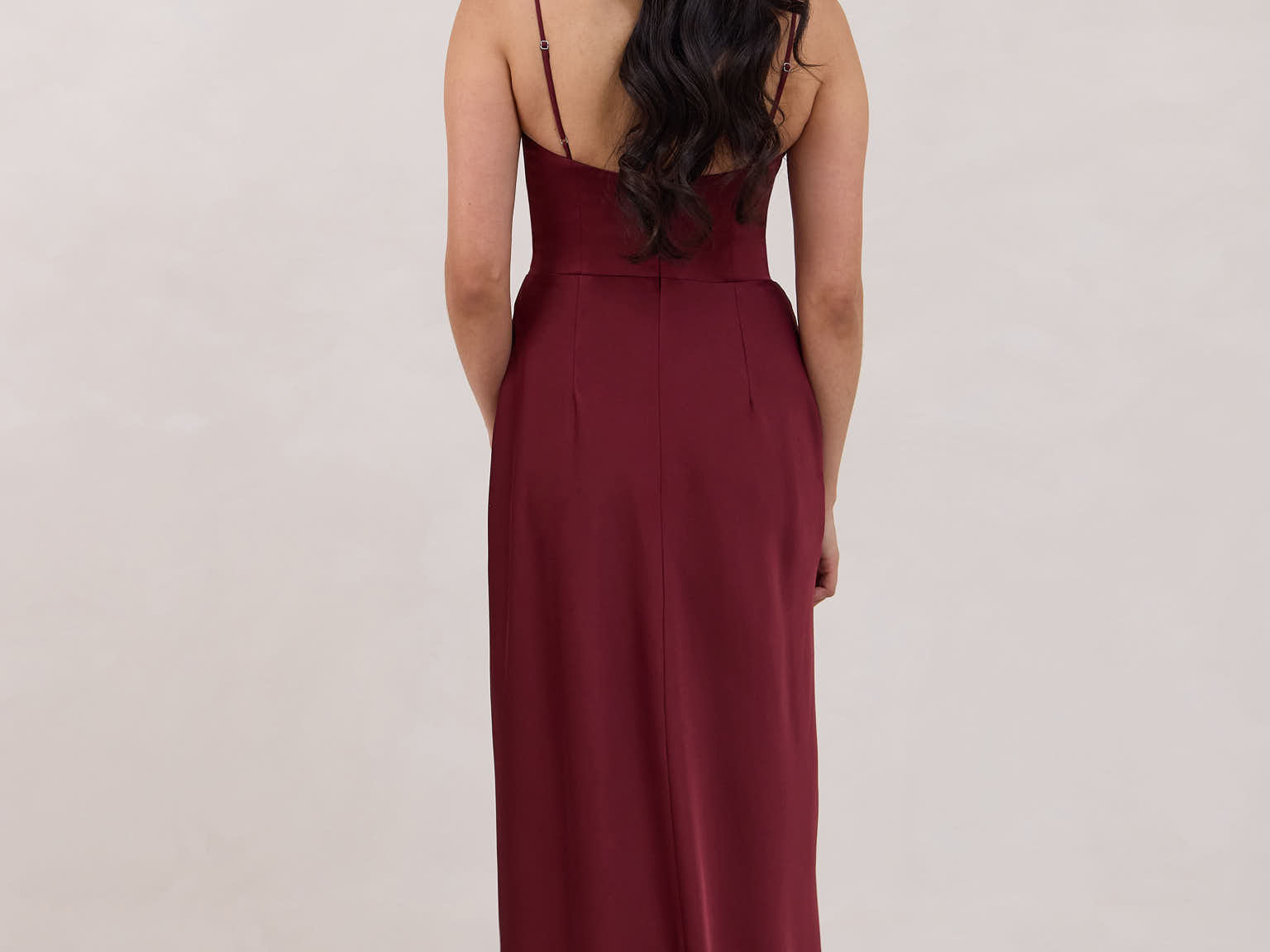 A woman is standing against a white backdrop facing the back wearing a burgundy, tea length satin bridesmaid dress. The dress has a boned bodice with a slight gathering at the bust, and a bias cut skirt that falls nicely over the hips. This is the Rosedale dress in merlot.