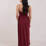 A woman is standing against a white backdrop facing the back wearing a burgundy, tea length satin bridesmaid dress. The dress has a boned bodice with a slight gathering at the bust, and a bias cut skirt that falls nicely over the hips. This is the Rosedale dress in merlot.