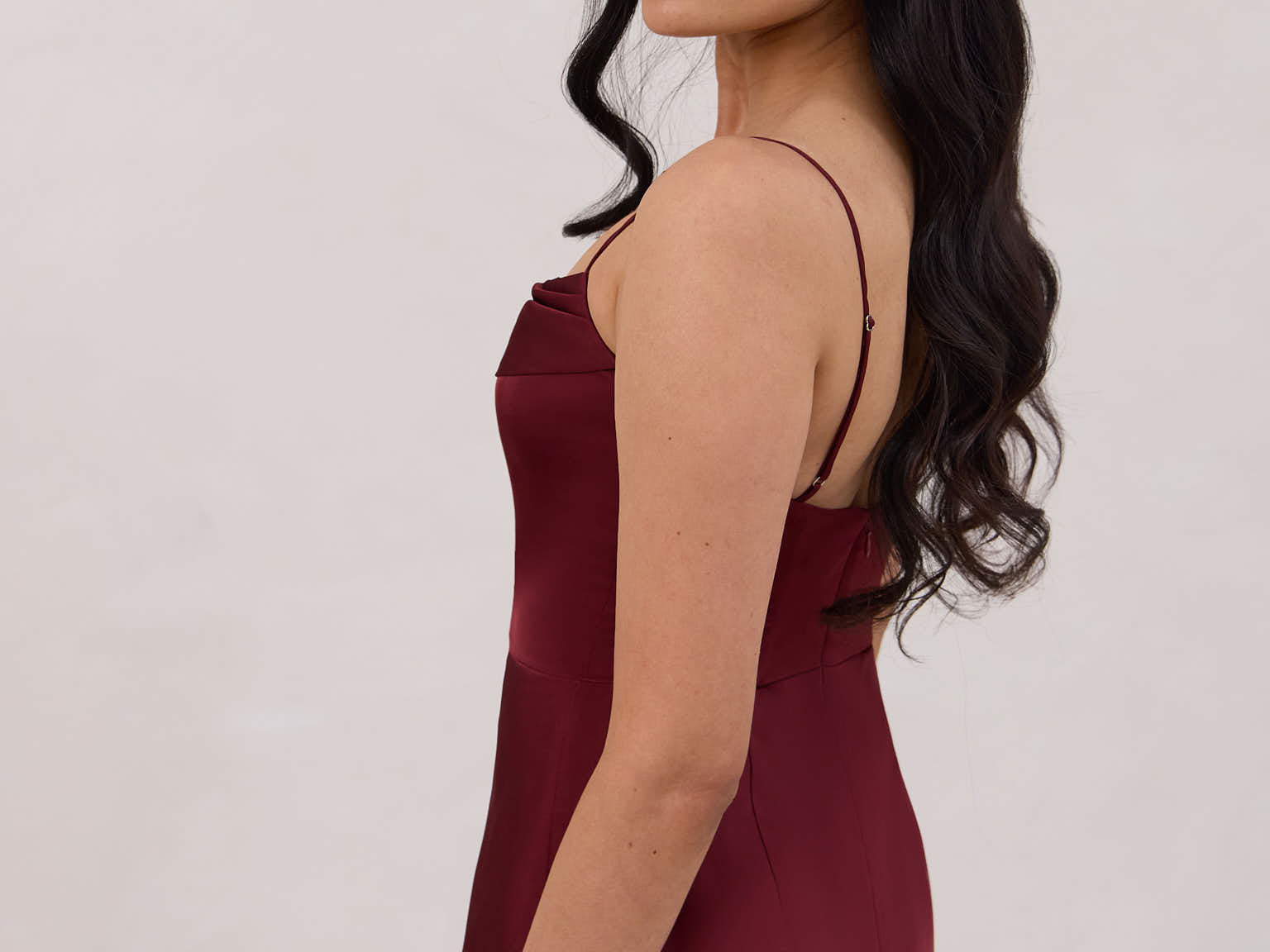 A woman is standing against a white backdrop wearing a burgundy, tea length satin bridesmaid dress. The dress has a boned bodice with a slight gathering at the bust, and a bias cut skirt that falls nicely over the hips. This is the Rosedale dress in merlot.