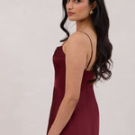 A woman is standing against a white backdrop wearing a burgundy, tea length satin bridesmaid dress. The dress has a boned bodice with a slight gathering at the bust, and a bias cut skirt that falls nicely over the hips. This is the Rosedale dress in merlot.