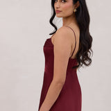 A woman is standing against a white backdrop wearing a burgundy, tea length satin bridesmaid dress. The dress has a boned bodice with a slight gathering at the bust, and a bias cut skirt that falls nicely over the hips. This is the Rosedale dress in merlot.