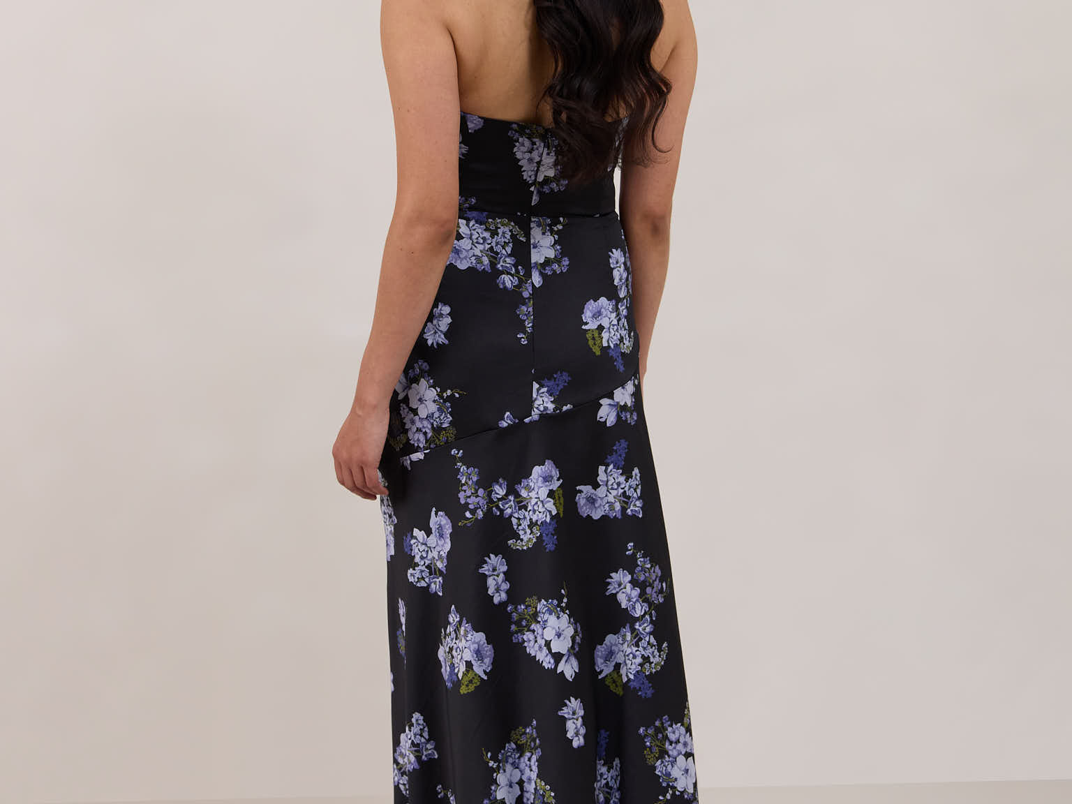 A woman is facing the back, wearing a dark blue strapless dress with a light blue floral print. The dress is fitted in the bodice and hip then flows into a fuller skirt. The dress is tea-length. This is the Sterling Dress in Viola.